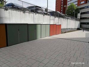 Mehrere farbige Gartenschränke an einer Wand auf einem gepflasterten Boden, mit geparkten Autos darüber.