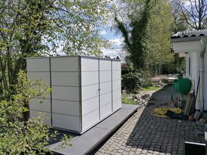 Ein weißer Schuppen neben einem Haus, umgeben von Bäumen und Gartenwerkzeug auf gepflastertem Weg.