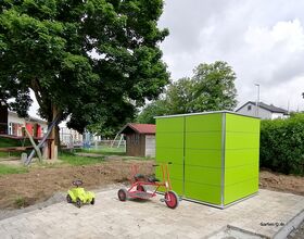 Ein grünes Gartenhaus neben Kinderspielzeug im Freien mit Bäumen und Häusern im Hintergrund.