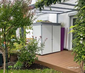 Gartenschrank mit einer Dachbegrünung auf einer Terrasse, grünem Rasen, Pflanzen und einem weißen Schuppen unter einer Pergola.