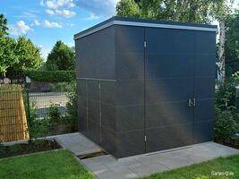 Ein moderner, dunkelgrauer Gartenschuppen steht auf gepflastertem Boden, umgeben von Grün und einem Zaun.