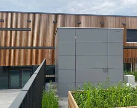 Ein modernes Gebäude mit Holzfassade, großem grauen Würfel und einem Holzpflanzkasten mit grünen Pflanzen im Vordergrund.