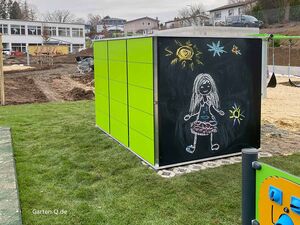 Grüner Schuppen mit Kreidezeichnungen: Sonne, Sterne und Kind auf Tafel. Baustelle und Schulgebäude im Hintergrund.