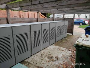 Metallene Gartenschränke unter einem Holzvordach, daneben ein Müllcontainer und parkende Autos im Hintergrund.