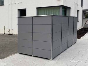 Ein großer, grauer Fahrradbox steht auf einer Terrasse vor einem modernen Gebäude.