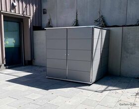 Ein grauer, moderner Gartenschrank steht auf einer Terrasse neben einer Hauswand.